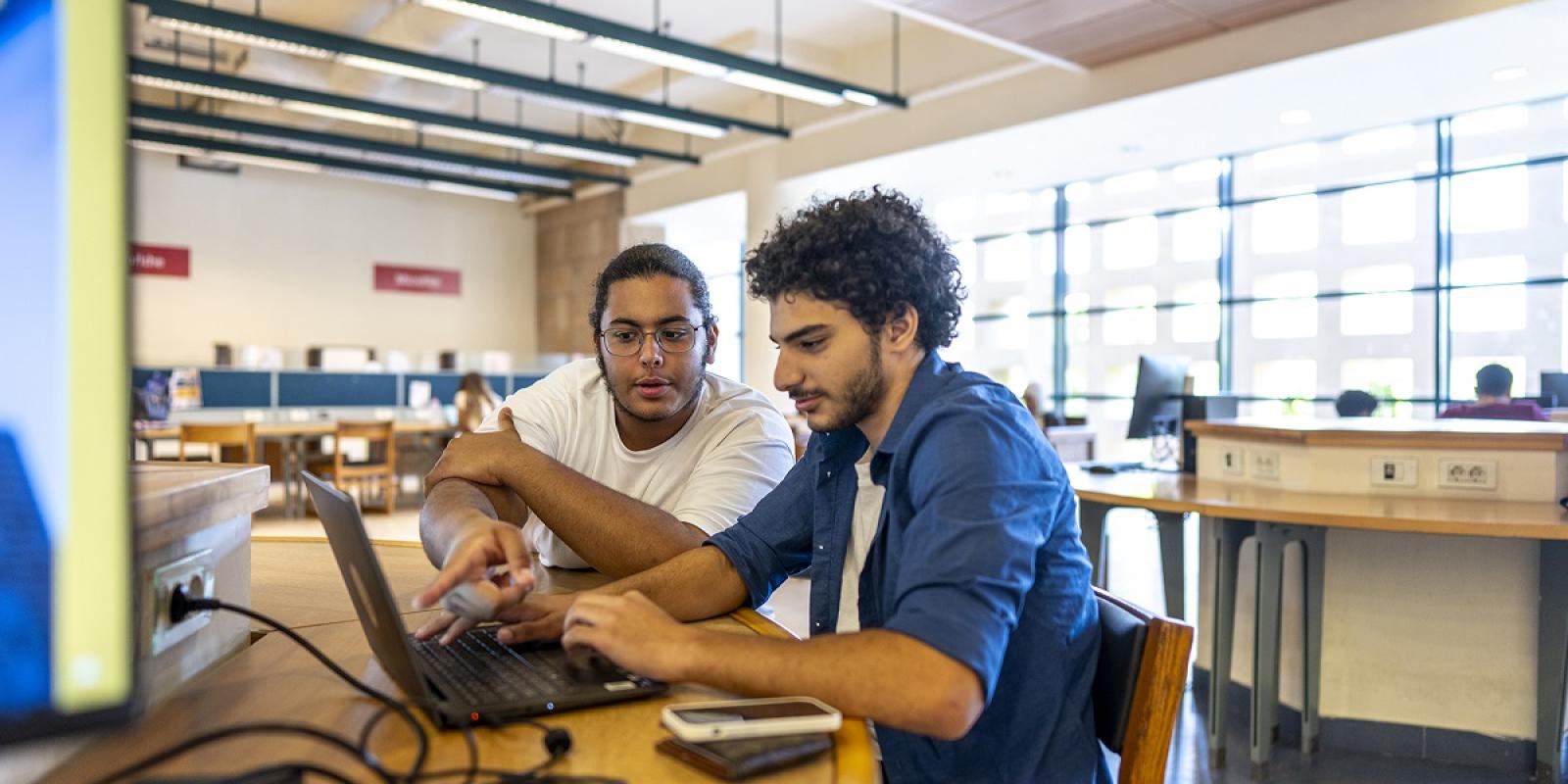 Two males are sitting at a desk in front of a laptop in a classroom