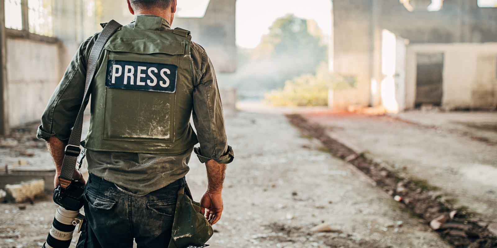 Man holding camera with a vest marked "press" walks in a damaged road full of rubble.