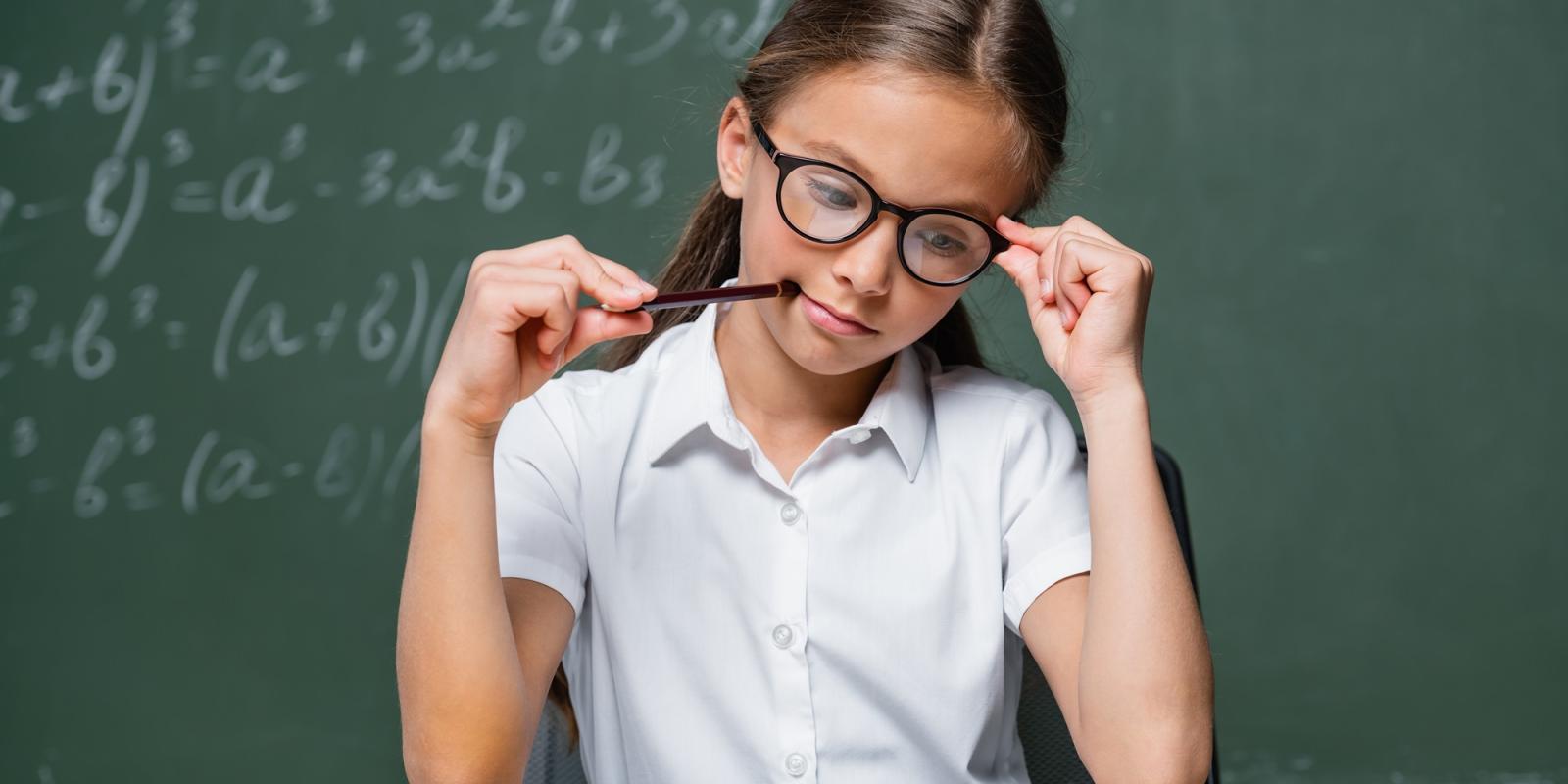 a young girl wearing a white tshirt and black eye glasses sitting on a desk holding a pen in her mouth