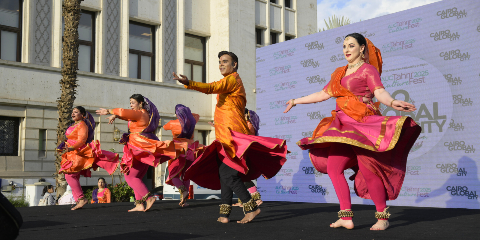 Dancers from Indian Embassy perform in colorful orange and pink saris on stage, arms spread