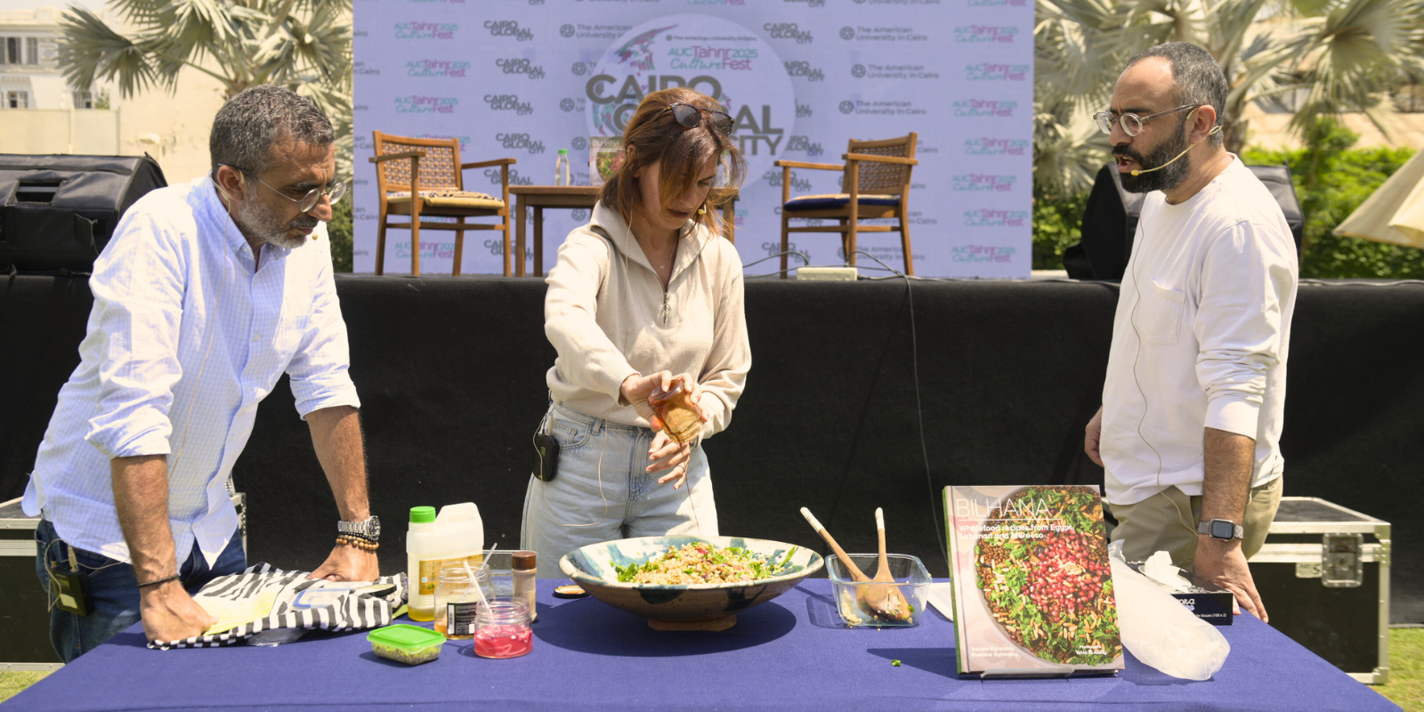 Yasmine (author) pours date molasses into a bowl of Freekeh with the photographer and moderator standing by in front of the stage at AUC Tahrir outdoors.
