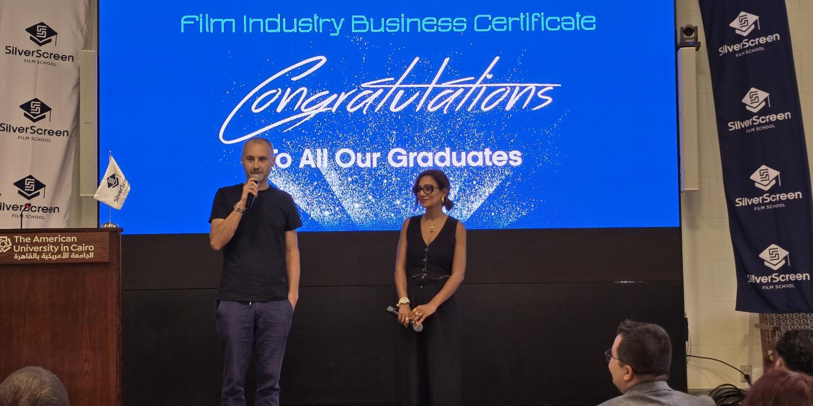 Co-founders of the Silver Screen Film School, a man and a woman, standing on stage speaking to the audience against a backdrop that says, "Congratulations to all our graduates"