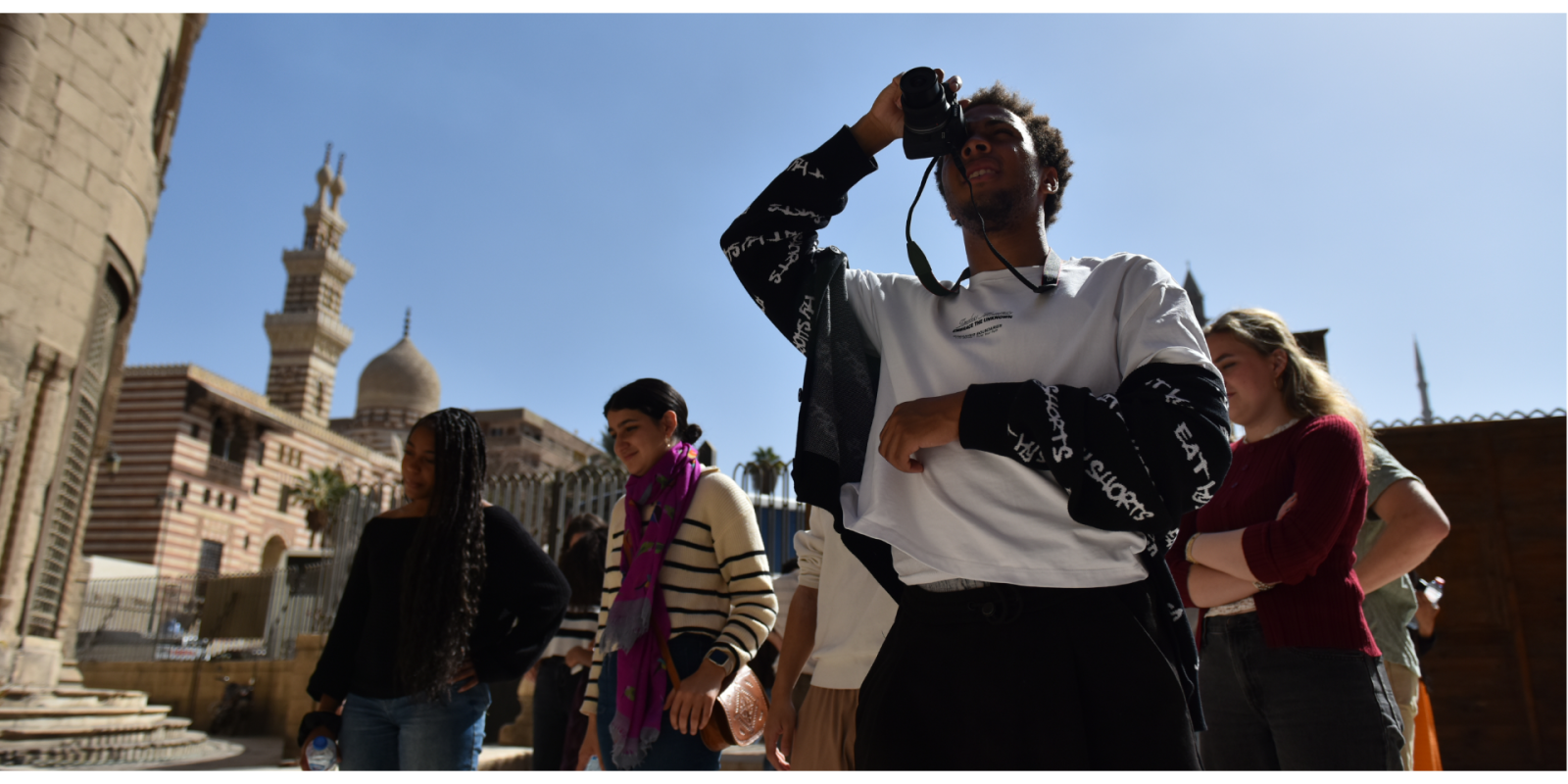 A student holds a camera to his eye while photographing something; a group of students stands behind him in conversation