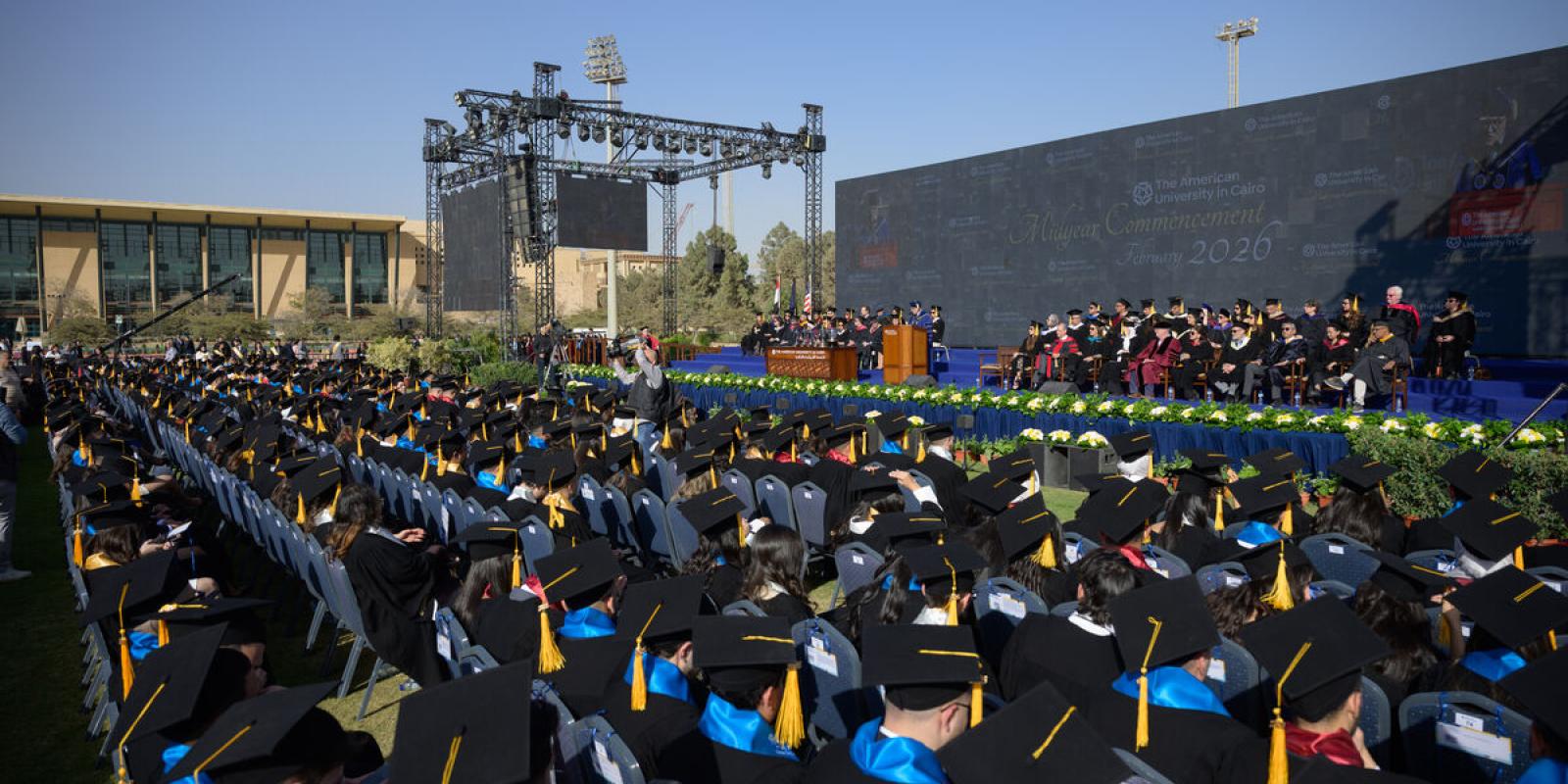 Wide outdoor view of AUC&rsquo;s midyear commencement ceremony, with rows of seated graduates facing a large stage and backdrop.
