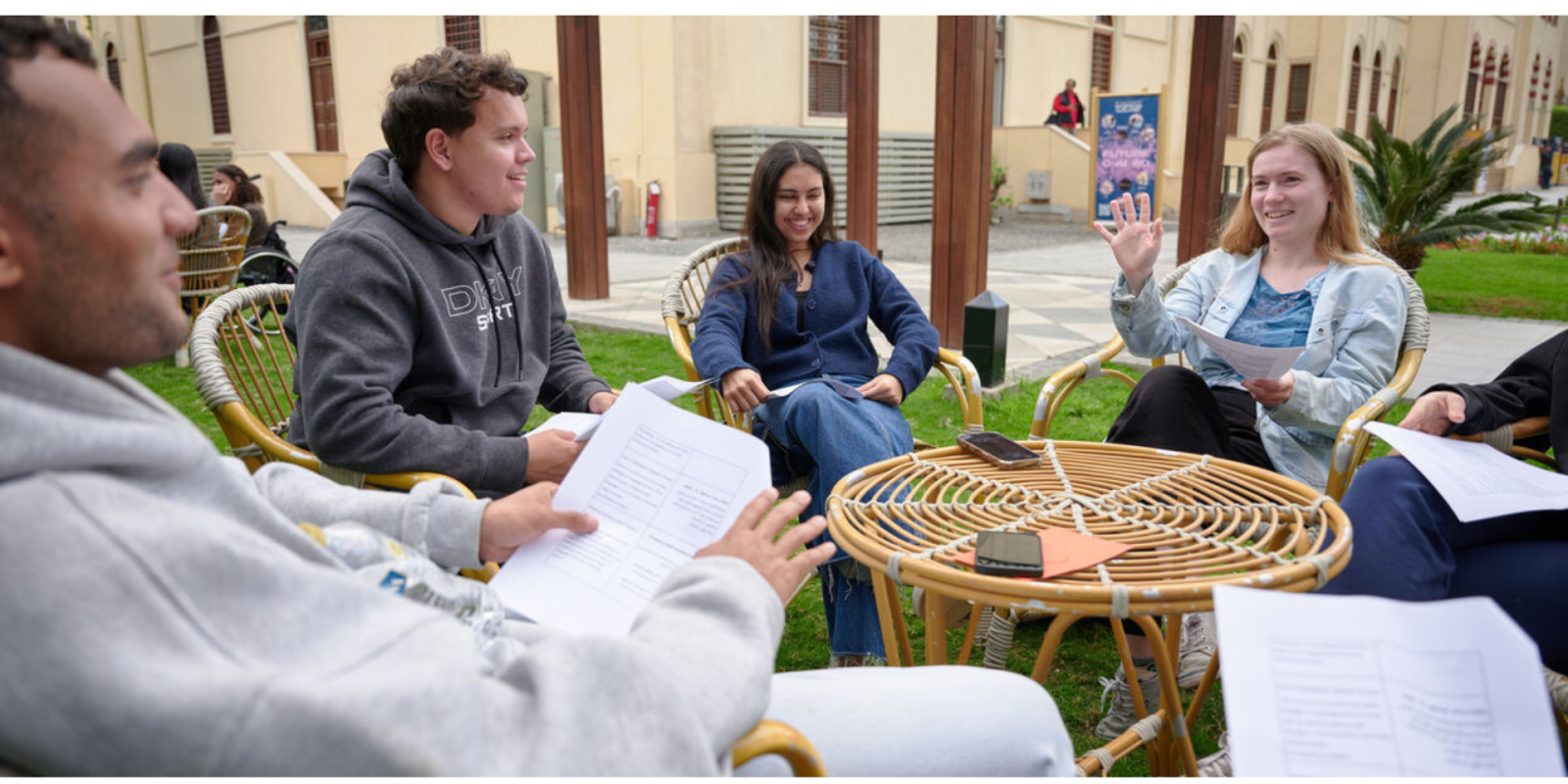 Students hold paper pamphlets while seated in chairs outside and talk with each other