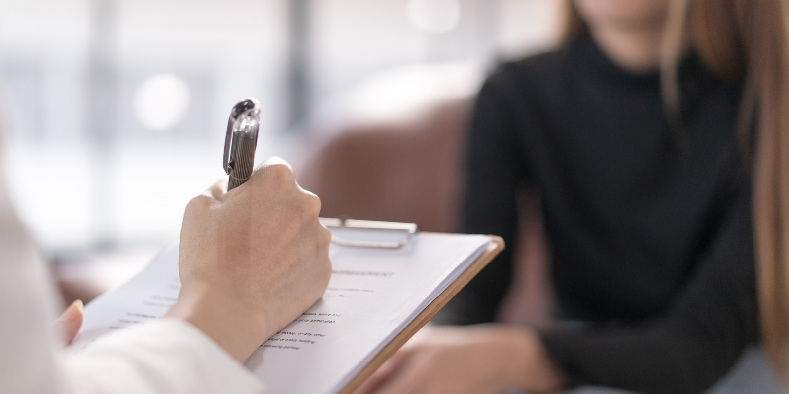 A hand of what appears to be a doctor writing on a paper and there is a person sitting across, explaining something using their hands