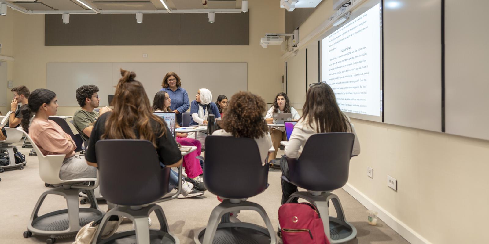 Students sit in a circle during a workshop discussion while an instructor stands nearby, with a projected document displayed on the wall.