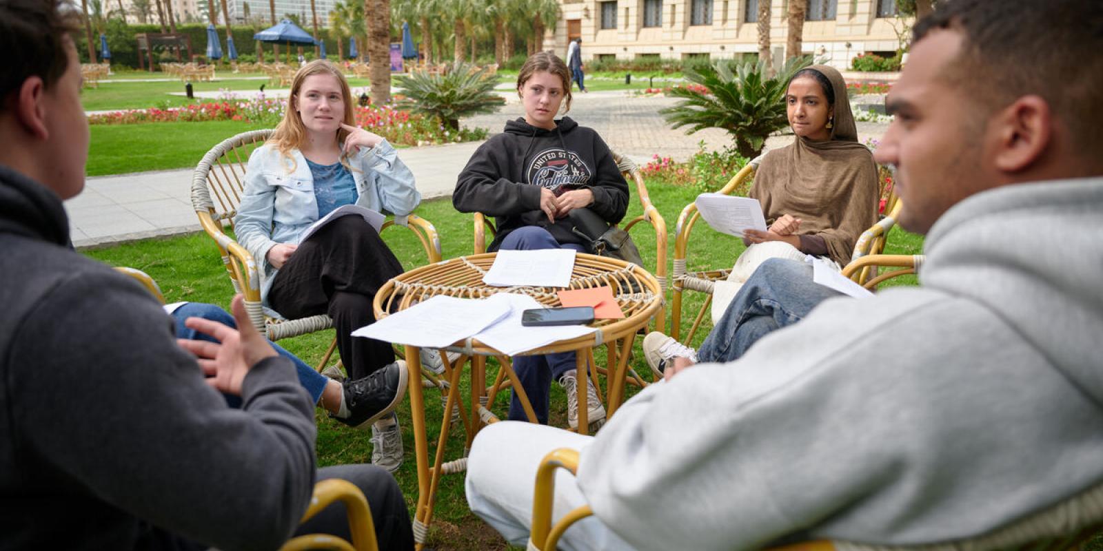 A group of students sit in a circle outside on campus holding papers while listening to a classmate speak during a discussion.