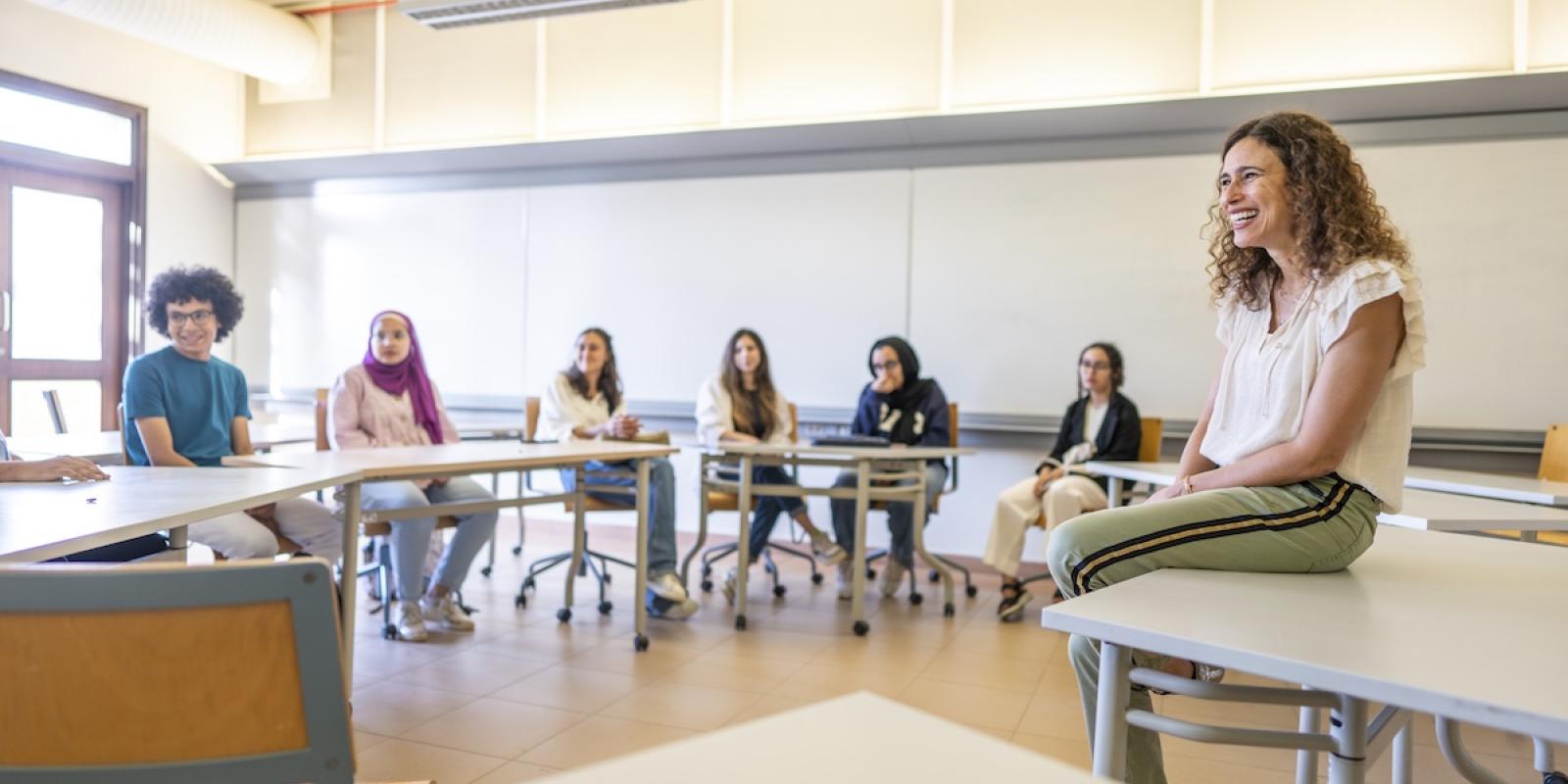 An instructor sits at the front of a classroom speaking with students seated in a circle during a discussion.