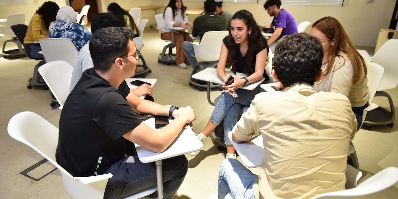 Students sit in small groups in a classroom discussing ideas and working together.