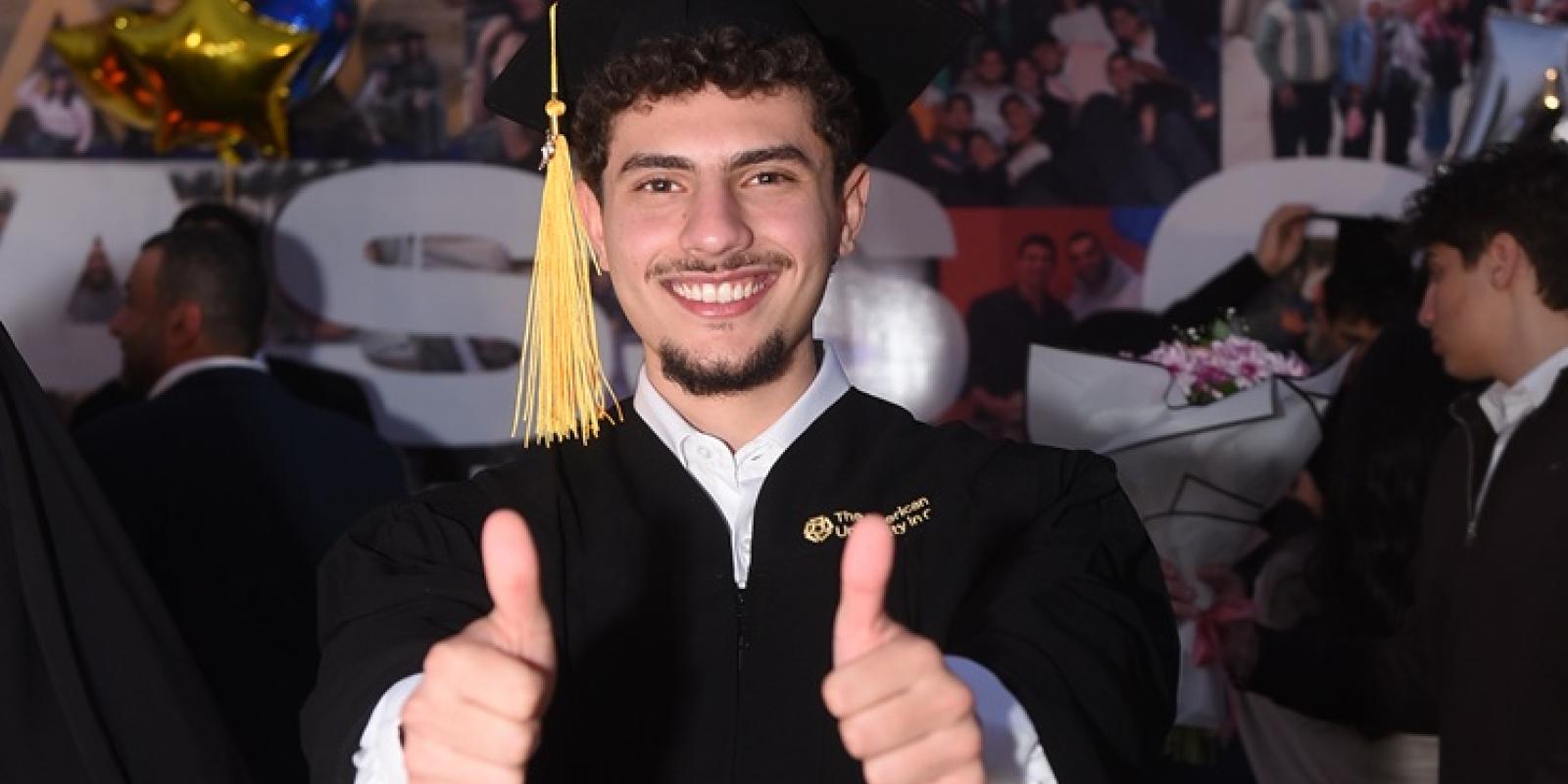 A male is standing and smiling. He is wearing his graduation capo and gown and giving two thumbs up