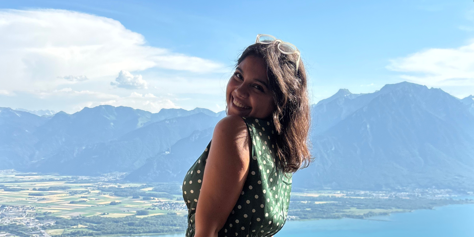 A girl standing in a balcony and behind her is a view of mountains, the sea, and greenery