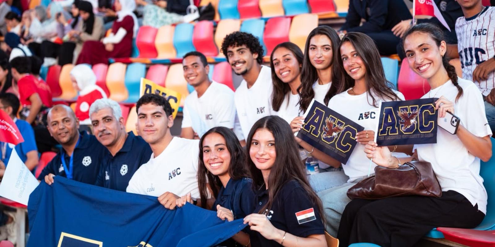 AUC students cheer from the stands holding an AUC banner. 