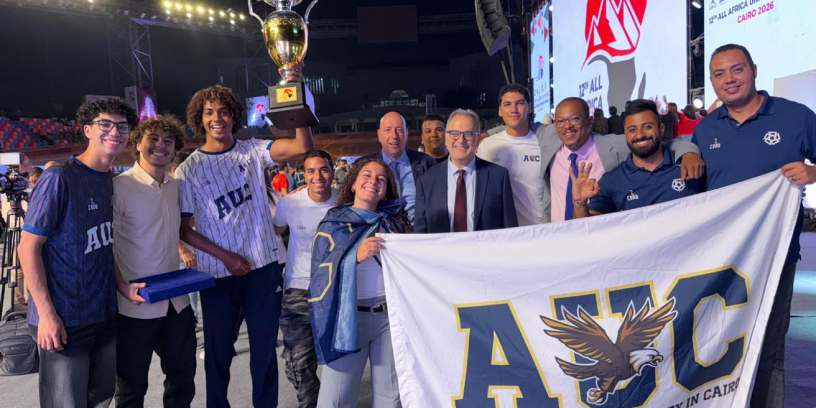 A group of AUC athletes stand with President Ahmad Dallal and Dean of Students Yorgen Marcel smiling, as one of them holds up the cup
