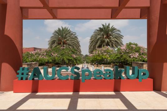 A photo of a campus statue of the words "#AUCSpeakUp" in blue on a red rectangular box. There are trees and the AUC garden in the background and a red veranda/pillars on the sides and over the top. 