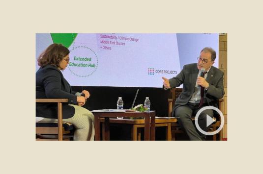 President Ahmad Dallal sits on a stage with moderator Amina Elbendary. He holds a microphone and gestures with his right hand, with a screen behind him showing an infographic explaining AUC's strategic priorities.