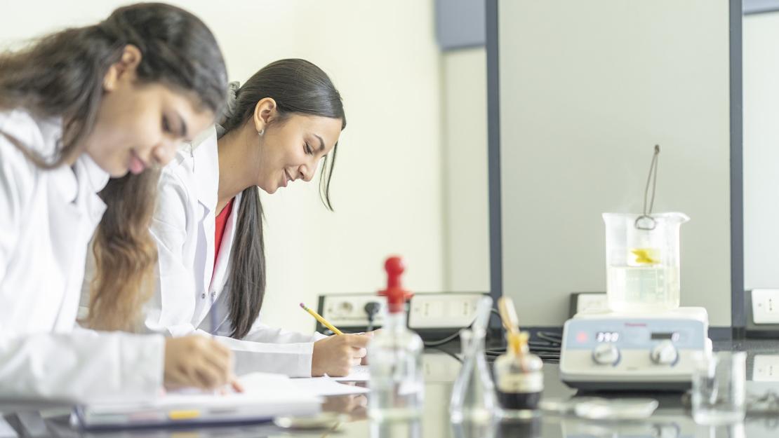 Two young women work on a counter in a science lab with glass beakers in front of them