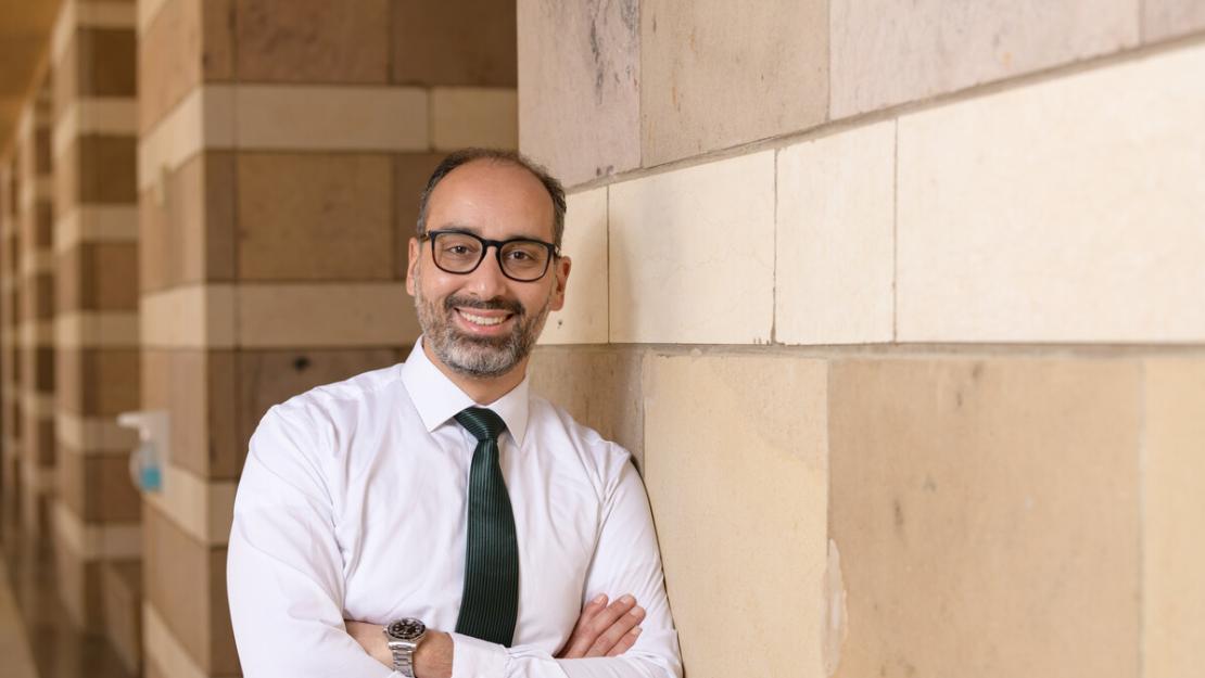 Smiling man in glasses wearing a button-up and blue tie leaning against a wall at AUC