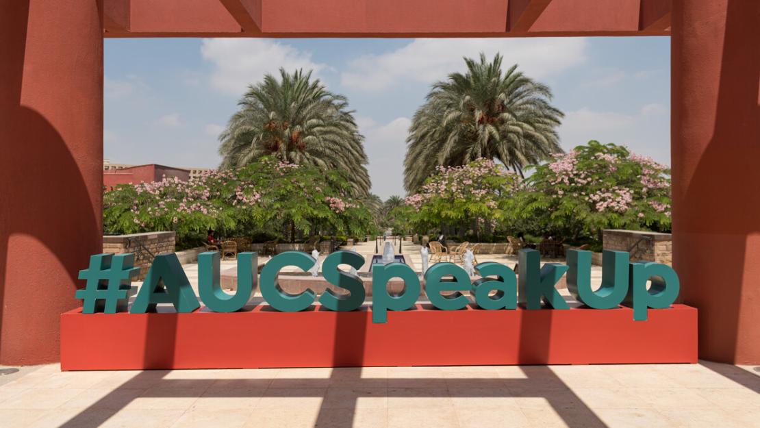 A photo of a campus statue of the words "#AUCSpeakUp" in blue on a red rectangular box. There are trees and the AUC garden in the background and a red veranda/pillars on the sides and over the top. 