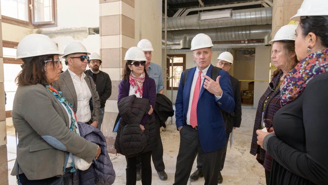 Board members and guests, men and women wearing suits and white hard hats visit construction site