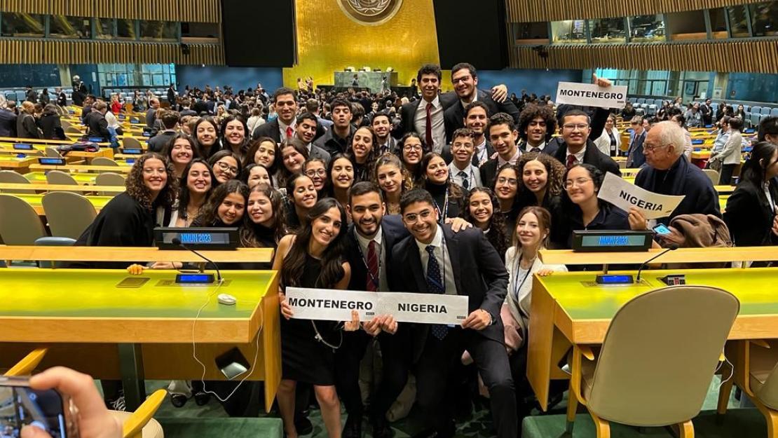 The CIMUN team stands wearing suits and professional dresses in an auditorium at the conference. Some students are holding signs saying "Montenegro" and "Nigeria"