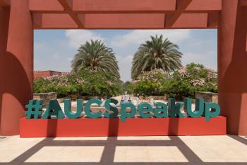 A photo of a campus statue of the words "#AUCSpeakUp" in blue on a red rectangular box. There are trees and the AUC garden in the background and a red veranda/pillars on the sides and over the top. 
