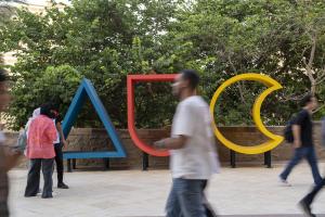 A large colorful wooden sign of AUC with students passing by