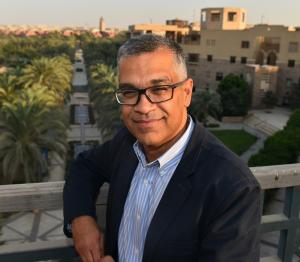 man wearing suit and glasses leans on a balcony edge overlooking auc gardens