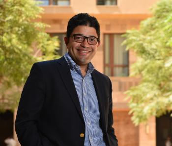 man wearing glasses smiles and stands in front of trees and a building