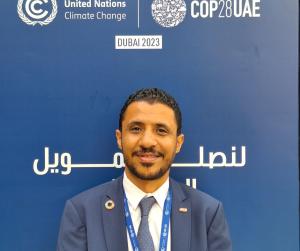 man in grey suit stands in front of a blue background with white COP28 logo on it