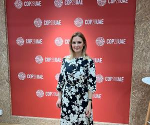 woman in a black and white dress stands in front of a red vinyl background containing the COP28 logo