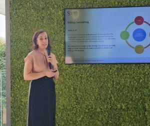 Woman wearing a black skirt and orange blouse stands in front of a green wall and presentation screen holding a microphone