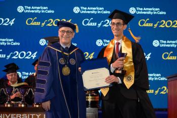 A man in a cap and gown stands next to the President of AUC holding a certificate and a golden trophy