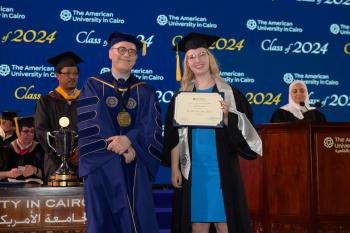 A woman stands in a cap and gown in front of a commencement background holding a certificate next to the AUC President.
