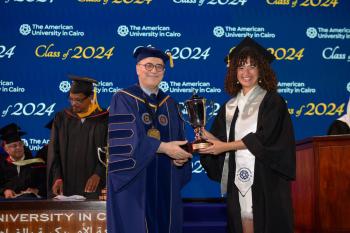 A woman in a cap and gown stands next to the President of AUC holding a certificate and a golden trophy