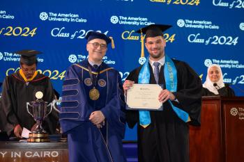 A man stands in a cap and gown in front of a commencement background holding a certificate next to the AUC President.
