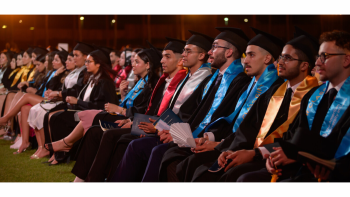 Students in caps and gowns sit in a row of chairs