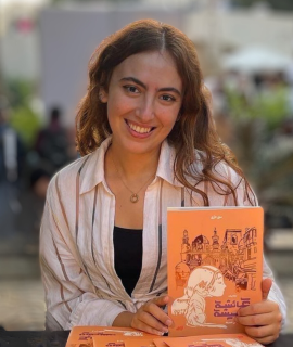 a girl with brown hair holding am orange book smiling