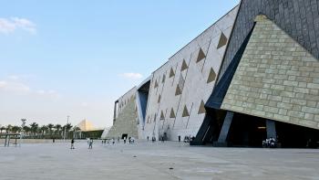 The Grand Egyptian Museum building from the outside with people walking in front of it and the pyramids in the background
