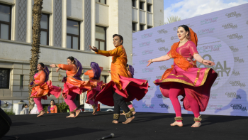 Dancers from Indian Embassy perform in colorful orange and pink saris on stage, arms spread