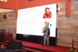A woman is talking into a microphone. There is a screen behind her that reads: Additional Scholarships