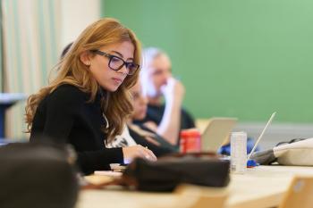 a girl with orange hair wearing black eye glasses and a black top sitting in a classroom