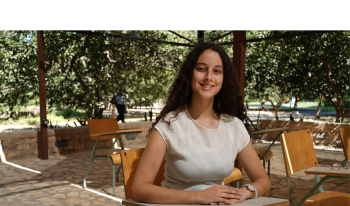 A student sits in a chair in an outside classroom surrounded by greenery.