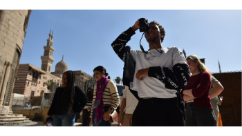 A student holds a camera to his eye while photographing something; a group of students stands behind him in conversation