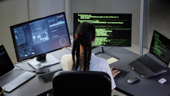 A women/girl sitting on a desk, with several desktops that has codes and programs open in front of her