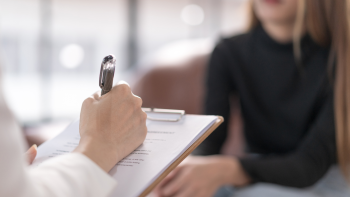A hand of what appears to be a doctor writing on a paper and there is a person sitting across, explaining something using their hands