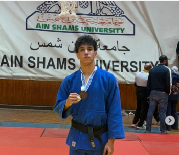 A male athlete is standing in a blue judo suit. He is holding a medal