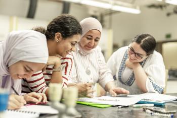 Four females collaborate at a table, smiling and reviewing notes and calculations together in a classroom setting.