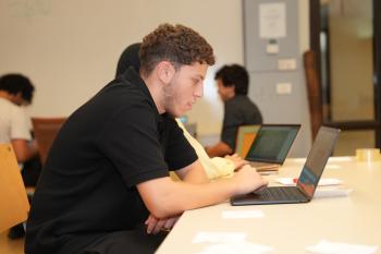 A student concentrates while typing on a laptop at a desk, with other students working in the background.