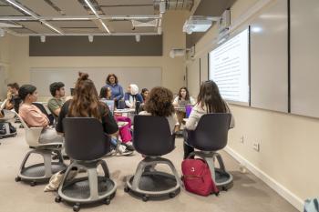 Students sit in a circle during a workshop discussion while an instructor stands nearby, with a projected document displayed on the wall.